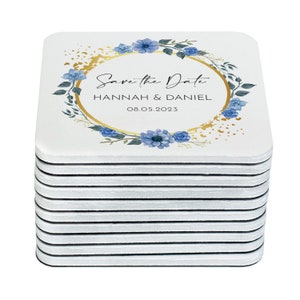 Puede incluir: Una pila de 15 posavasos cuadrados blancos con un diseño floral azul y dorado. Los posavasos presentan el texto "Save the Date" con los nombres "Hannah & Daniel" y la fecha "08.05.2023".