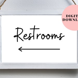 Könnte beinhalten: Ein weißer gerahmter Schild mit schwarzem Text, der "Restrooms" lautet, mit einem schwarzen Pfeil, der nach links zeigt. Das Schild hängt an einer weißen Wand.
