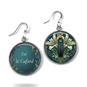 Peut inclure: Boucles d'oreilles rondes avec crochets argentés. L'une porte l'inscription "J'ai le Cafard" sur fond turquoise, ornée de fleurs. L'autre présente un insecte noir entouré de fleurs blanches et de feuillage.
