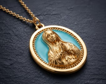 May include: A gold-toned oval pendant necklace featuring a raised relief of a figure with hands clasped in prayer. The figure is set against a bright turquoise enamel background, framed by a beaded gold border, and attached to a delicate gold chain.