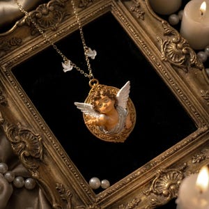 Puede incluir: Collar dorado con colgante de querub&iacute;n. El querub&iacute;n tiene alas blancas y est&aacute; enmarcado en un &oacute;valo. El collar se exhibe sobre una superficie negra, con un espejo dorado y perlas en el fondo. Un dise&ntilde;o cl&aacute;sico y elegante.