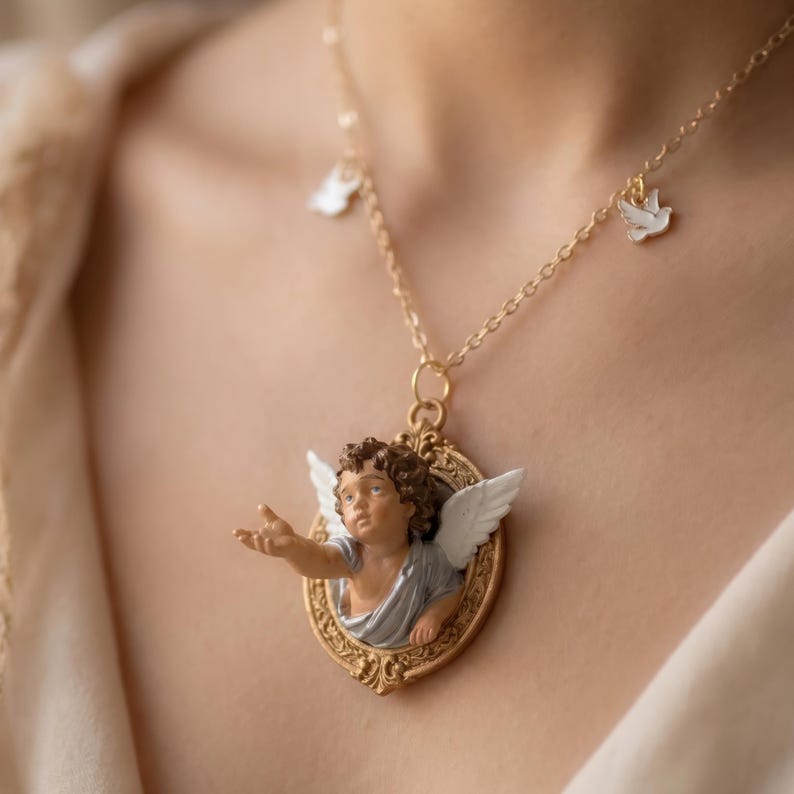 Puede incluir: Collar dorado con un colgante de querub&iacute;n. El querub&iacute;n tiene cabello casta&ntilde;o y alas blancas, enmarcado en un marco dorado. El collar tambi&eacute;n incluye peque&ntilde;os dijes de paloma blancos.
