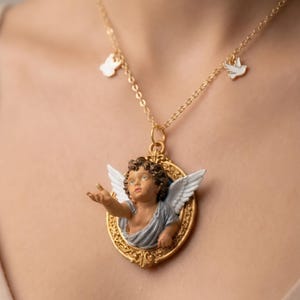 Puede incluir: Collar dorado con un colgante con una figura de querub&iacute;n en un marco ovalado dorado. El querub&iacute;n tiene alas blancas y viste una prenda azul gris&aacute;ceo. El collar tambi&eacute;n tiene peque&ntilde;os dijes de una paloma y una forma.
