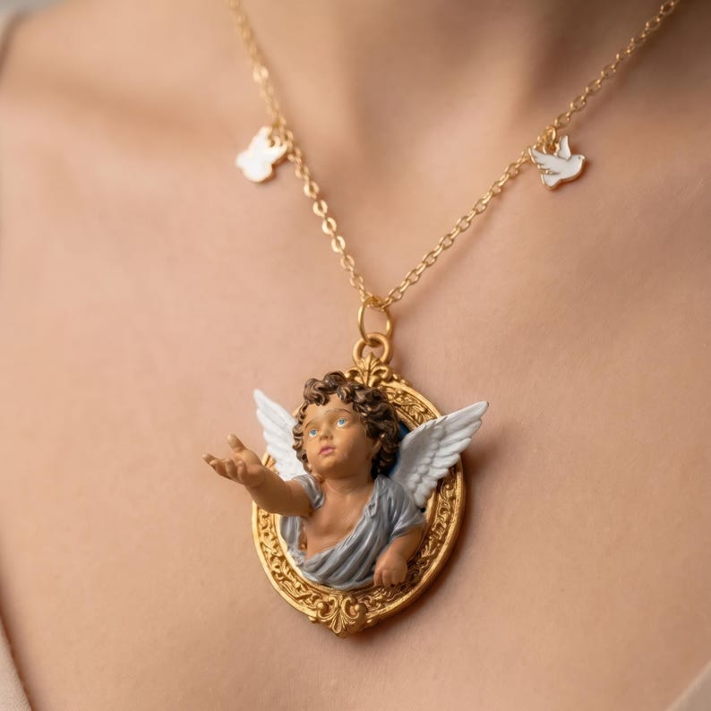 Puede incluir: Collar dorado con un colgante con una figura de querub&iacute;n en un marco ovalado dorado. El querub&iacute;n tiene alas blancas y viste una prenda azul gris&aacute;ceo. El collar tambi&eacute;n tiene peque&ntilde;os dijes de una paloma y una forma.