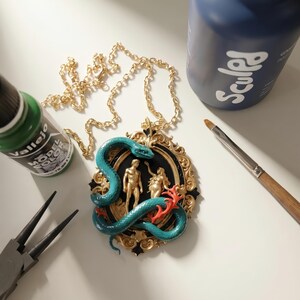 May include: A gold-colored chain necklace with a pendant featuring a teal snake coiled around gold-colored figures. The pendant has a black background and a gold-colored ornate frame. Paint bottles and tools are in the background.