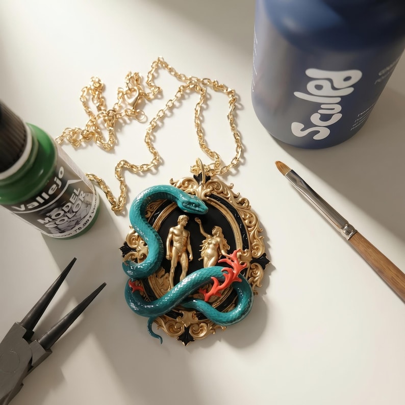 May include: A gold-colored chain necklace with a pendant featuring a teal snake coiled around gold-colored figures. The pendant has a black background and a gold-colored ornate frame. Paint bottles and tools are in the background.