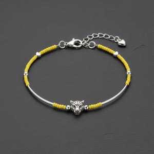 Peut inclure: Un bracelet argenté avec un fil de perles jaunes et un charm en forme de tête de léopard argenté. Le bracelet est doté d'une chaîne délicate avec un détail en forme de cœur. Fond sombre.