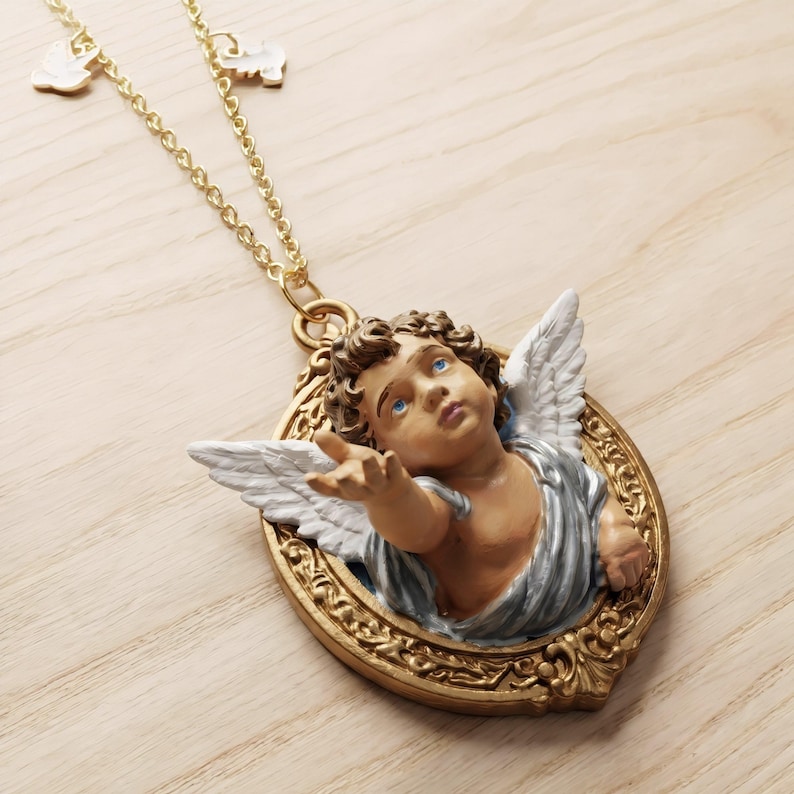 Puede incluir: Collar dorado con colgante de querub&iacute;n. El colgante muestra un querub&iacute;n con ojos azules, cabello casta&ntilde;o y alas blancas, en un marco dorado. La cadena tiene peque&ntilde;os dijes. El colgante mide aproximadamente 5 cm de alto.