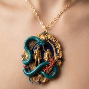 May include: A gold-toned pendant necklace featuring a detailed scene with figures and a serpent. The pendant has a blue background, gold frame, and a teal snake wrapping around the figures. The figures are gold-colored.