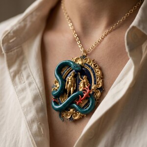 May include: Gold-toned pendant necklace featuring a detailed design. The pendant showcases a blue and gold oval frame with a central scene of two figures and a green snake. Red coral-like accents add contrast. The necklace is worn against a cream-colored shirt.