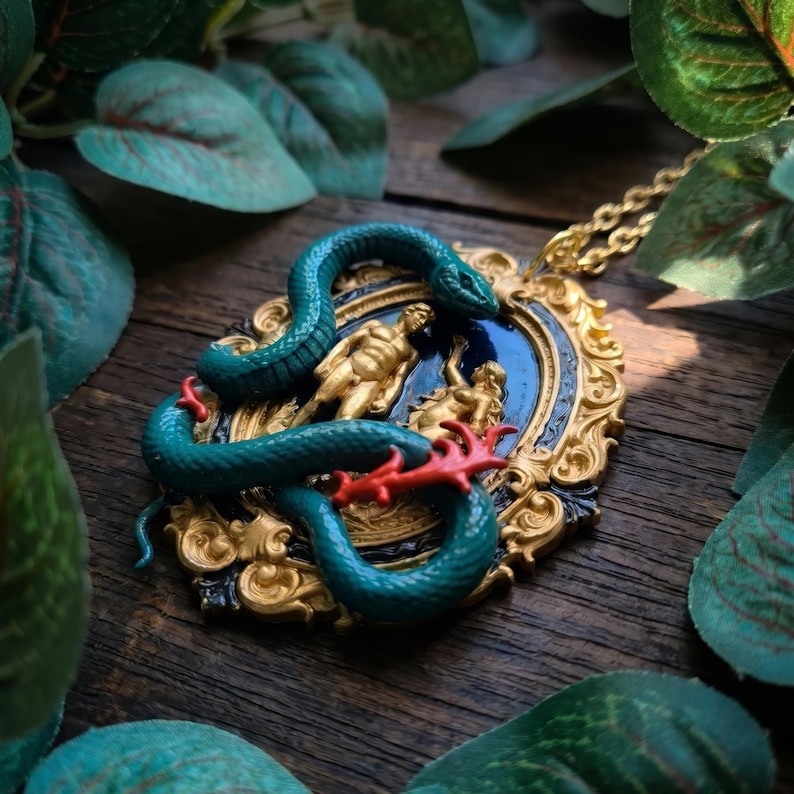 May include: A teal snake pendant necklace with a gold chain. The pendant features a gold-colored frame with a blue center, and a gold-colored relief of two figures. The snake is teal with red accents. The necklace is on a wooden surface.