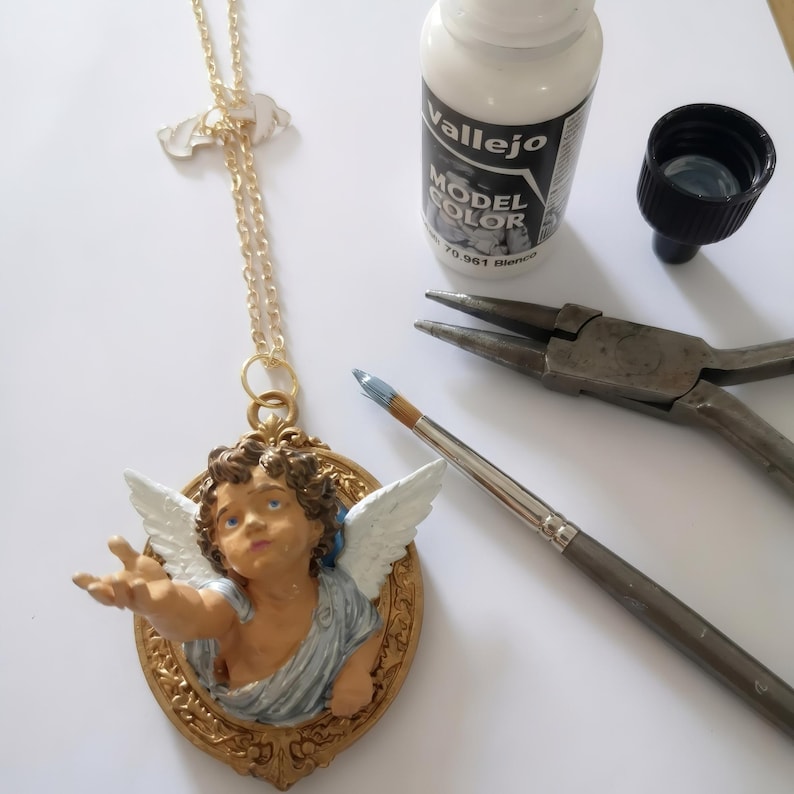 Puede incluir: Collar con colgante dorado con un querub&iacute;n con el brazo extendido y alas blancas. El querub&iacute;n est&aacute; dentro de un marco ovalado dorado. Tambi&eacute;n se ven un bote de pintura blanca, un pincel y unos alicates.