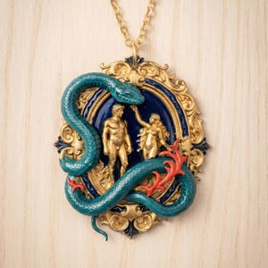 May include: A gold-toned pendant necklace featuring a teal snake coiled around a framed scene of two figures. The frame has a blue background and gold accents. Red coral-like details add contrast.
