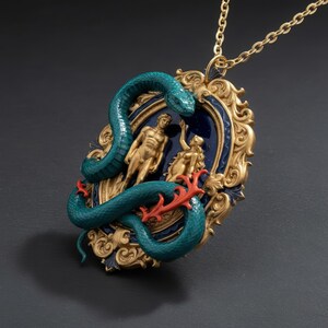 May include: A gold-toned pendant necklace featuring a teal snake coiled around a framed scene of two figures. The frame is gold with a dark blue background. A red coral-like element is also present.