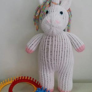 以下が含まれることがあります： 虹色のたてがみと角を持つ白いニットのユニコーンのおもちゃ。ユニコーンは、カラフルなプラスチック製の編み機3つのある白い表面の上に立っています。