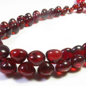 May include: A strand of deep red, translucent, heart-shaped garnet beads arranged in a curved formation. The polished stones exhibit a smooth, rounded surface with a glossy finish, reflecting light across their dark crimson, faceted-like appearance.