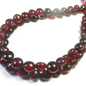 May include: A strand of deep red, translucent, round beads with a smooth, polished finish. The beads are strung together in a continuous loop, showing a rich, dark crimson hue that catches the light, with a bright red cord visible at the top.