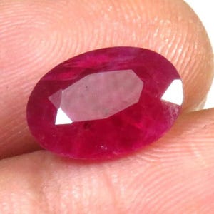 Puede incluir: Gema de rubí de talla ovalada con un tono rojo intenso. La gema tiene múltiples facetas que reflejan la luz, creando un efecto brillante. El rubí se sostiene en una mano, mostrando su tamaño y color.
