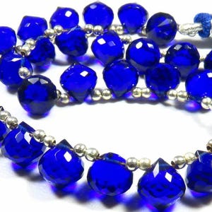 Peut inclure: Un collier de perles à facettes en forme de cœur, d'un bleu profond, alternant avec de petites perles intercalaires rondes de couleur argentée. Le collier est disposé en courbe et comporte un pompon en fil bleu avec un embout argenté.