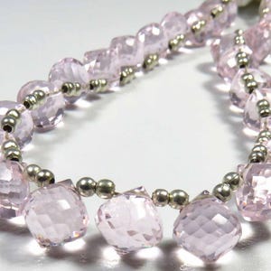 Può includere: Una fila di perline sfaccettate di colore rosa chiaro a forma quadrata, alternate a piccole perline distanziatrici rotonde di colore argento metallizzato. Le perline sono disposte in una linea curva, mostrando una superficie traslucida.