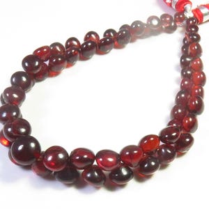 Pode incluir: Um fio de contas de pedra preciosa vermelho-escuras, translúcidas e polidas, em forma de pequenas gotas arredondadas. As contas estão enfiadas num laço, presas no topo por uma borla de tecido vermelho vivo com detalhes em metal prateado.
