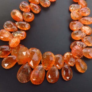 May include: A collection of faceted, pear-shaped sunstone beads in vibrant shades of orange and gold. Each gemstone displays a translucent quality with internal shimmering inclusions, arranged in a curved formation against a dark background.