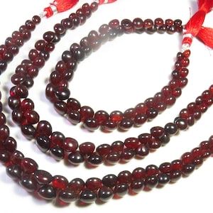 May include: Three strands of deep red, smooth, teardrop-shaped beads arranged in a nested curve. Each strand is finished with a bright red tassel attached by a silver-toned metal cap, creating a uniform look across the collection of beaded jewellery.