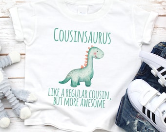 May include: A white short-sleeve t-shirt featuring a green illustration of a dinosaur with orange spikes. The text reads "COUSINSAURUS" above the dinosaur and "LIKE A REGULAR COUSIN, BUT MORE AWESOME" below it in green lettering.