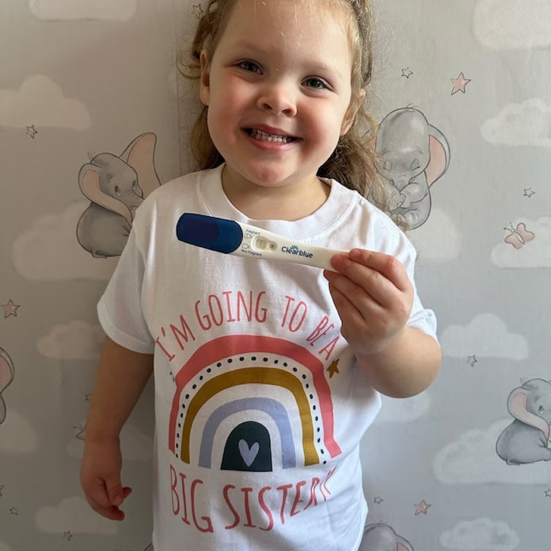 A happy little girl with curly hair, holding up a positive pregnancy test, wearing a white 'I'M GOING TO BE A BIG SISTER' t-shirt with a colorful rainbow above the announcement text, standing against a whimsical wallpaper