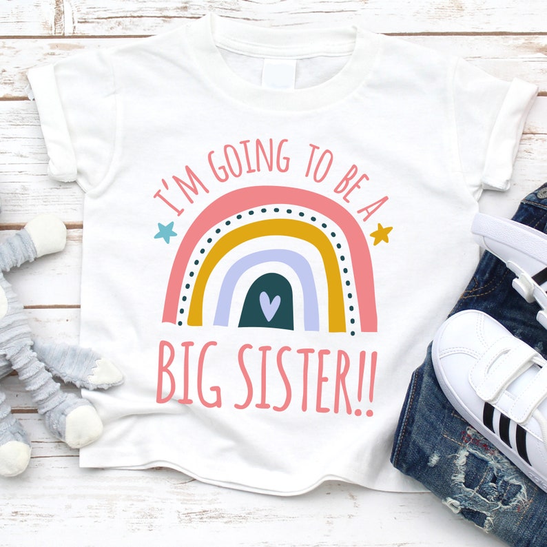 White 'BIG SISTER!!' t-shirt with a vibrant rainbow graphic above the announcement text, displayed with casual children's jeans and white sneakers on a rustic wooden background.