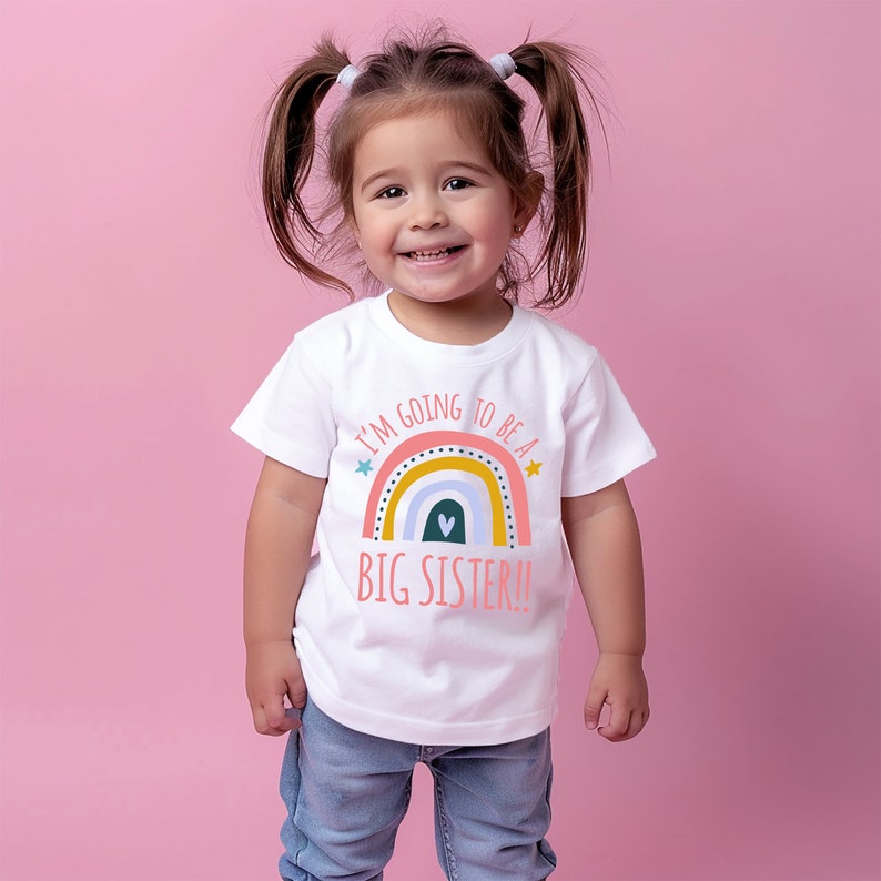 Cheerful toddler girl with pigtails smiling and wearing a white 'BIG SISTER!!' t-shirt with a colorful rainbow and heart graphic, standing against a soft pink background.