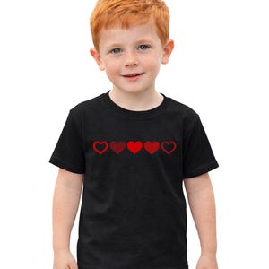 Peut inclure: T-shirt noir à manches courtes orné d'une rangée de cinq cœurs rouges sur la poitrine. Les cœurs ont des motifs variés, certains pleins et d'autres soulignés. Le t-shirt est porté par un jeune enfant aux cheveux roux et à la peau claire.