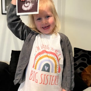 Excited blonde toddler proudly holding up a sonogram picture above her head, wearing a layered white 'I'M GOING TO BE A BIG SISTER!!' t-shirt with a rainbow design, in a cozy living room setting.