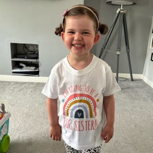 A joyful toddler with hair in pigtail buns, grinning widely and wearing a white t-shirt that reads 'I'M GOING TO BE A BIG SISTER!!' with a colorful rainbow graphic, in a bright living room setting.
