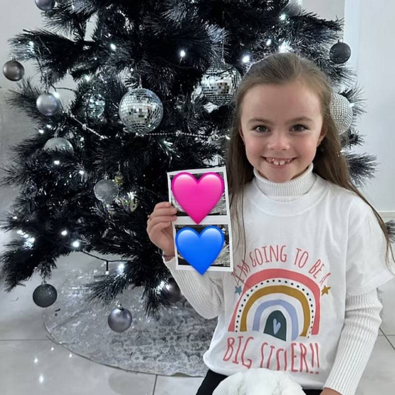 "A smiling girl standing in front of a Christmas tree, wearing a white 'I'M GOING TO BE A BIG SISTER!!' t-shirt with a rainbow design, holding a sonogram photo covered by heart emojis for a holiday-themed pregnancy announcement.