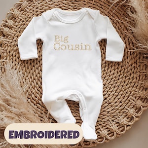 May include: White long-sleeved baby grow with the words "Big Cousin" embroidered in gold on the front.