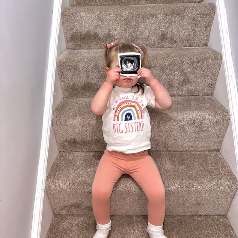 A toddler girl sitting on the stairs, holding up a sonogram photo in front of her face, wearing a 'BIG SISTER!' t-shirt with a colorful rainbow and heart design, in a playful and heartwarming pregnancy announcement scene.
