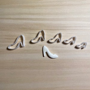 May include: A collection of white high heel shoe-shaped cookie cutters in various sizes. One solid shoe is present, along with several outline cutters. The cutters are arranged on a light brown wooden surface.