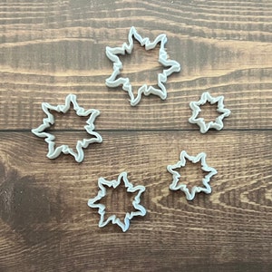May include: Five white plastic snowflake cookie cutters of varying sizes. The cookie cutters are arranged on a wooden surface.