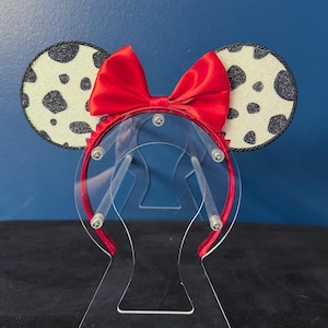 May include: A pair of mouse ears with a red satin bow and a headband. The ears are white with black spots and a glittery texture, outlined with black trim. The headband is red.