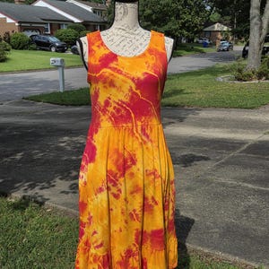 May include: A sleeveless knee-length dress featuring a vibrant tie-dye pattern in shades of bright orange and deep magenta. The garment has a round neckline, a fitted bodice, and a gathered skirt section that ends in a ruffled hemline detail.