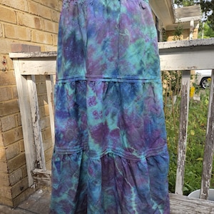May include: A long, tiered skirt featuring a tie-dye pattern in shades of deep purple, teal, and blue. The skirt has an elasticized waistband with gathered fabric and two horizontal stitched seams that create distinct tiers along the length.
