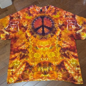 May include: A short-sleeve t-shirt featuring a vibrant tie-dye pattern in shades of orange, yellow, and deep red. The center of the shirt displays a prominent dark purple peace sign graphic, surrounded by intricate, swirling dye effects.