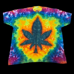 May include: A short-sleeve t-shirt featuring a vibrant tie-dye pattern in shades of yellow, orange, blue, purple, and green. The center of the shirt displays a large, dark, solid-colored leaf-shaped graphic set against a bright yellow background.