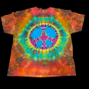 May include: A short-sleeve t-shirt featuring a vibrant tie-dye pattern with a prominent central peace sign graphic. The design incorporates concentric circles of bright blue, magenta, yellow, and green, surrounded by a mottled blend of orange and red.