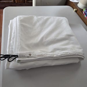 May include: A folded white fabric tarp featuring reinforced edges with metal grommets and black elastic bungee cords attached to the corner. The material appears smooth and opaque, neatly stacked in a square shape with visible stitching along the borders.