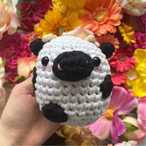 Könnte beinhalten: Ein weißer gehäkelter Kuh-Plüsch mit schwarzen Augen, einer schwarzen Nase und schwarzen Flecken. Der Plüsch sitzt in einem Bett aus bunten Blumen.