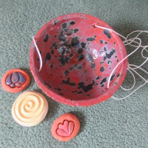 May include: A red ceramic hanging planter with black speckles and white string. Three small, decorative ceramic pieces in orange and cream colors are next to the planter. One piece has a spiral design, and the other two have floral designs.