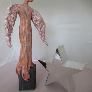 Op de afbeelding: Een keramisch engelbeeld met een getextureerd, bruin lichaam en witte vleugels. De engel staat op een zwarte basis. Een zilverkleurig stervormig object bevindt zich op de voorgrond.