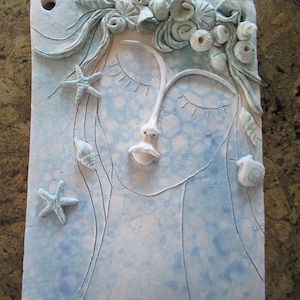 Puede incluir: Una baldosa de cerámica azul claro con un rostro esculpido con los ojos cerrados, adornado con conchas marinas y estrellas de mar. La obra de arte tiene una superficie texturizada y un agujero para colgar. El diseño evoca un tema costero.